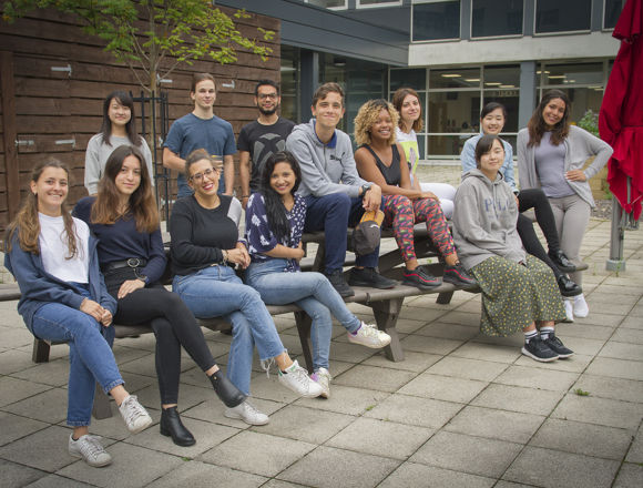 Group of students sitting on a bench, posing for a photo outside a college campus building.